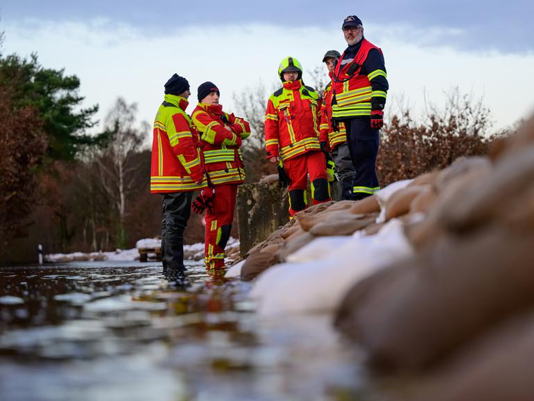 Mehrere ehrenamtliche Einsatzkräfte der Feuerwehr in Uniform stehen zur Besprechung in einer Gruppe zusammen neben aufeinandergeschichteten Sandsäcken.