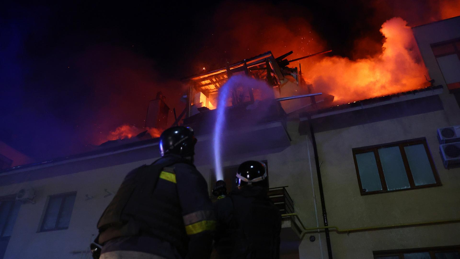 Es ist dunkel. Zwei Feuerwehrleute spritzen mit einem Schlauch Wasser in ein brennendes oberes Stockwerk eines Hauses, aus dem Flammen schlagen. Es ist dunkel. Zwei Feuerwehrleute spritzen mit einem Schlauch Wasser in ein brennendes oberes Stockwerk eines Hauses, aus dem Flammen schlagen.