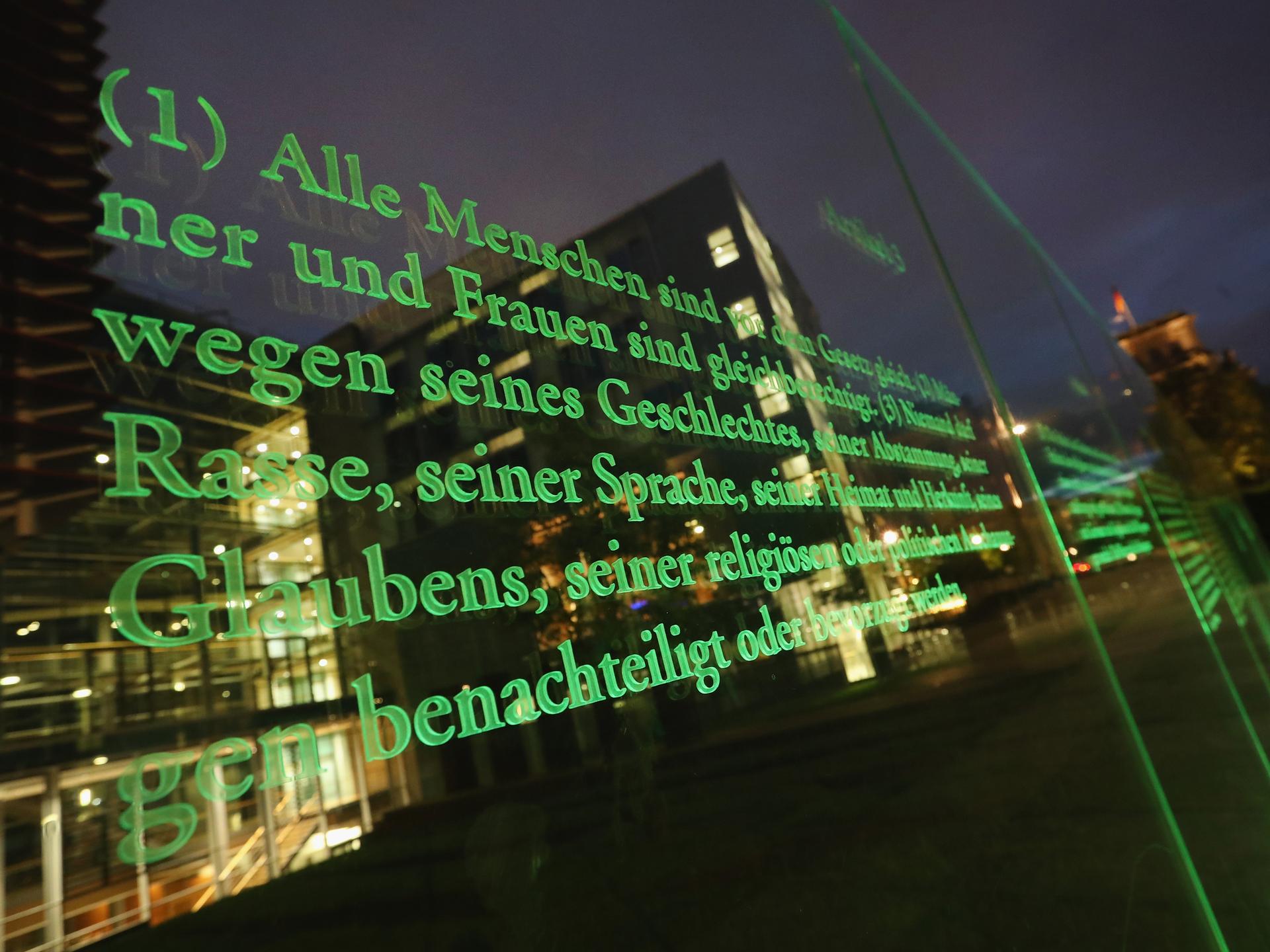 Ein Artikel des deutschen Grundgesetzes auf einer Glastafel am Reichstagsufer, Spiegelungen von Gebäuden und Lichtern.