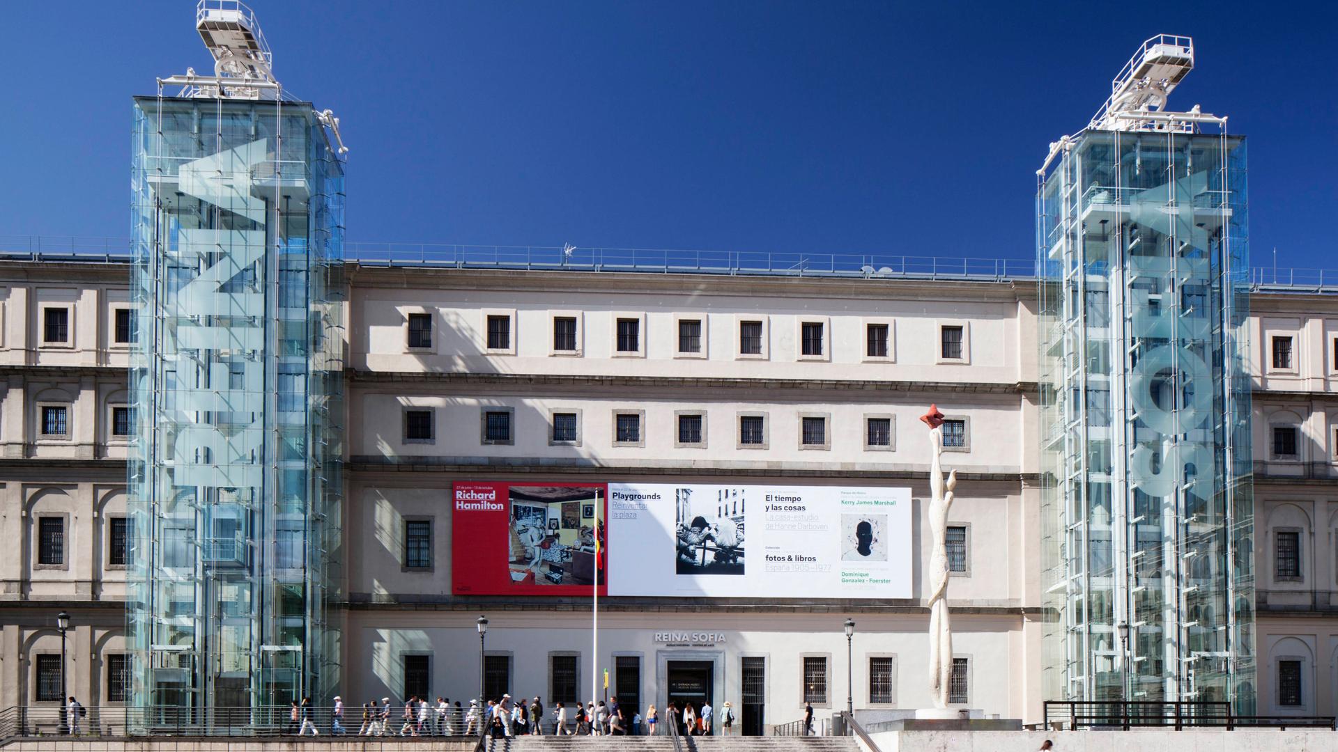 Außenansicht eines Gebäudes und der Haupteingangsfassade des Museo Nacional Centro de Arte Reina Sofia in Madrid, vor dem sich Menschen zwischen den beiden verglasten Türmen mit den Aufzügen und Treppen tummeln.