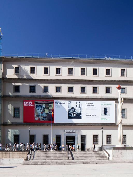 Außenansicht eines Gebäudes und der Haupteingangsfassade des Museo Nacional Centro de Arte Reina Sofia in Madrid, vor dem sich Menschen zwischen den beiden verglasten Türmen mit den Aufzügen und Treppen tummeln.
