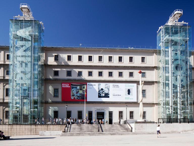 Außenansicht eines Gebäudes und der Haupteingangsfassade des Museo Nacional Centro de Arte Reina Sofia in Madrid, vor dem sich Menschen zwischen den beiden verglasten Türmen mit den Aufzügen und Treppen tummeln.