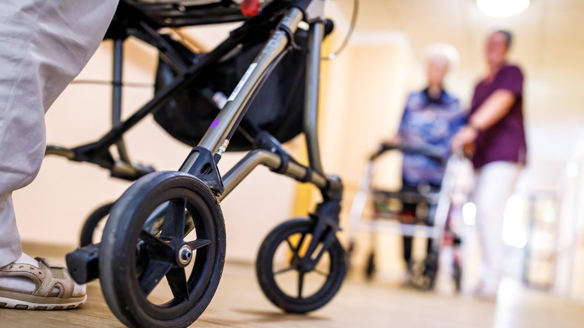 Senioren mit Rollator begegnen sich auf dem Flur. Von der vorderen Person links im Bild sind die Beine und der Rollator zu sehen, rechts unscharf und weiter hinten geht eine Seniorin in Begleitung einer Pflegekraft.