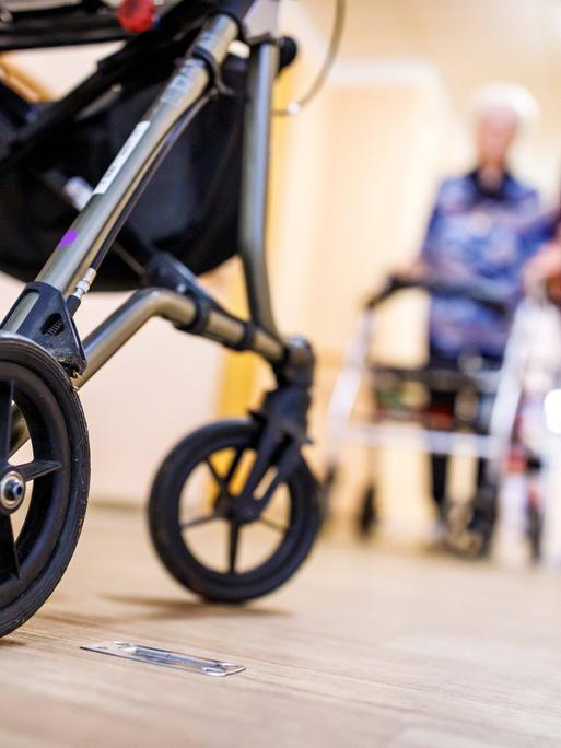 Senioren mit Rollator begegnen sich auf dem Flur. Von der vorderen Person links im Bild sind die Beine und der Rollator zu sehen, rechts unscharf und weiter hinten geht eine Seniorin in Begleitung einer Pflegekraft. Senioren mit Rollator begegnen sich auf dem Flur. Von der vorderen Person links im Bild sind die Beine und der Rollator zu sehen, rechts unscharf und weiter hinten geht eine Seniorin in Begleitung einer Pflegekraft.