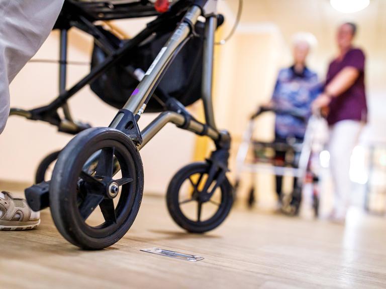 Senioren mit Rollator begegnen sich auf dem Flur. Von der vorderen Person links im Bild sind die Beine und der Rollator zu sehen, rechts unscharf und weiter hinten geht eine Seniorin in Begleitung einer Pflegekraft. Senioren mit Rollator begegnen sich auf dem Flur. Von der vorderen Person links im Bild sind die Beine und der Rollator zu sehen, rechts unscharf und weiter hinten geht eine Seniorin in Begleitung einer Pflegekraft.