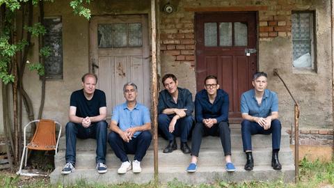 Fünf Männer sitzen in Jeans auf den Stufen eines Hauseinganges, das sich in einem romantisch verwildertem Zustand befindet.
