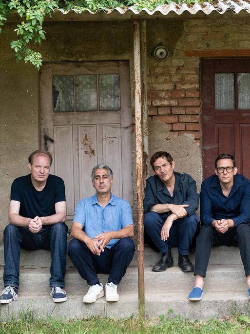 Fünf Männer sitzen in Jeans auf den Stufen eines Hauseinganges, das sich in einem romantisch verwildertem Zustand befindet.