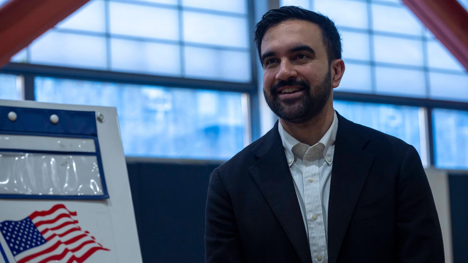 Zohran Mamdani steht in einer Halle. Er lächelt in Richtung der Kamera. 