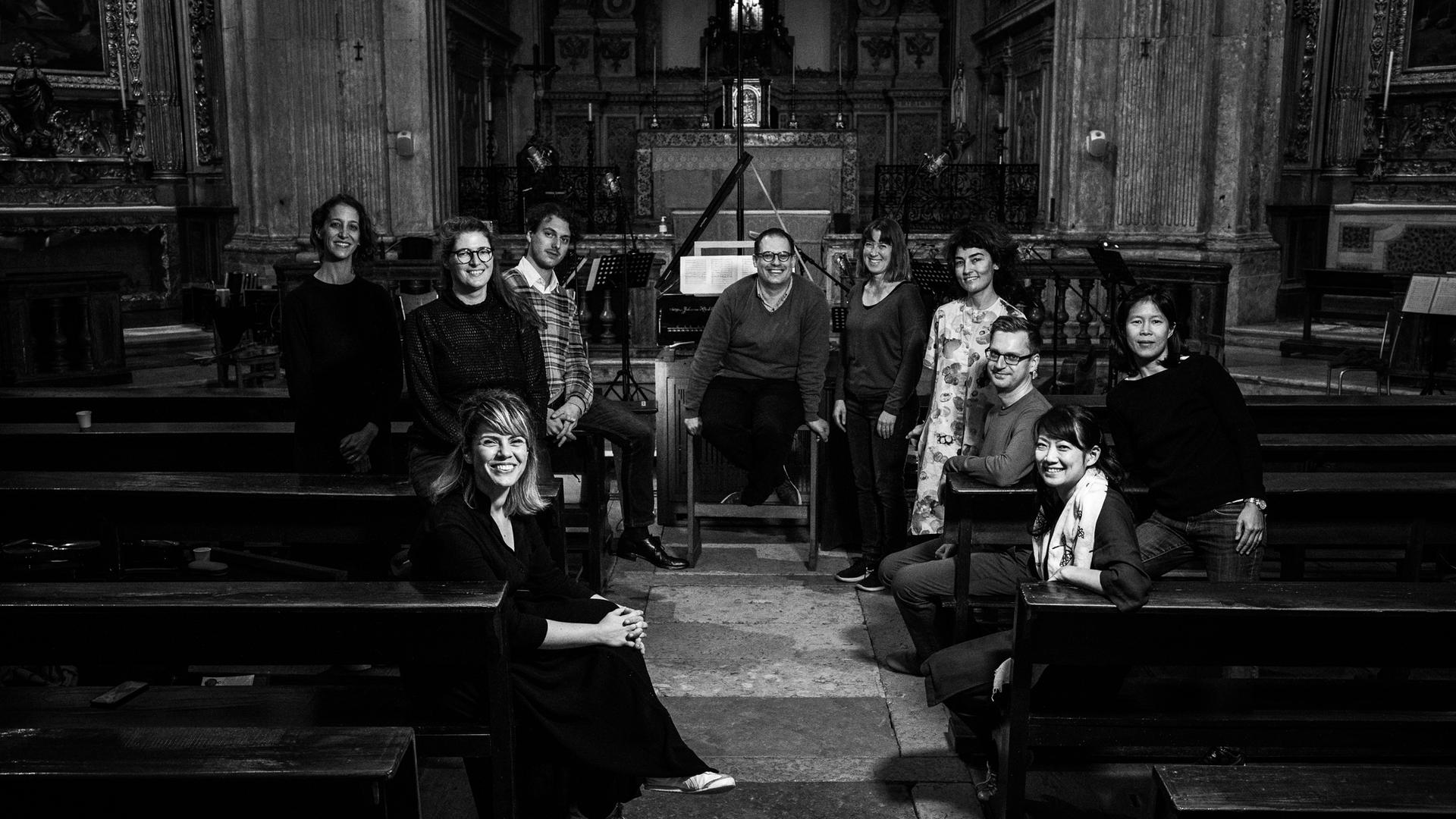 Zehn Menschen stehen in einer Kirche vor einem Altarraum, zum Teil sitzen sie auf den Kirchenbänken. Alle lächeln. Das Bild ist in schwarz-weiß-Tönen gehalten.