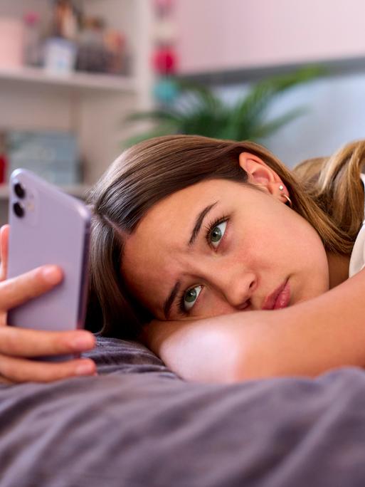 Eine Jugendliche liegt auf dem Bett. Sie schaut traurig auf ihr Handy.