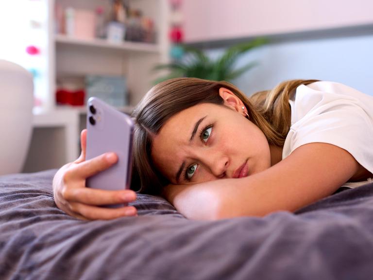 Eine Jugendliche liegt auf dem Bett. Sie schaut traurig auf ihr Handy.