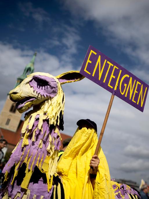 Bei einer Demonstration hält eine Person, die in gelbe Tücher gehüllt ist und auf einem Pappmaschee-Alpaka sitzt, ein Schild in die Luft auf dem "Enteignen" steht.