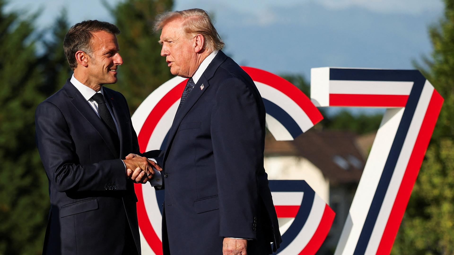 Der französische Präsident Emmanuel Macron (l.) begrüßt US-Präsident Donald Trump während des G7-Gipfels. Im Hintergrund ist das Logo des G7-Gipfels, eine überdimensional große Kombination aus G und 7 aufgestellt. Das Logo ist blau-weiß-rot gestreift. Der französische Präsident Emmanuel Macron (l.) begrüßt US-Präsident Donald Trump während des G7-Gipfels. Im Hintergrund ist das Logo des G7-Gipfels, eine überdimensional große Kombination aus G und 7 aufgestellt. Das Logo ist blau-weiß-rot gestreift.