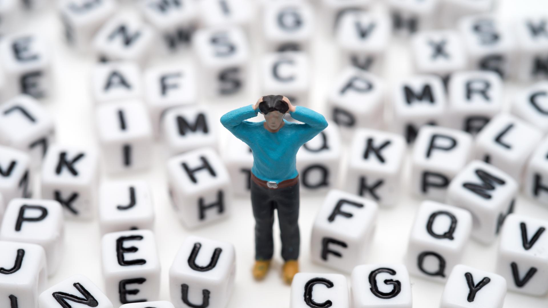 Eine kleine Spielfigur steht, die Hände an den Kopf gehoben, zwischen lauter Buchstabenwürfeln 
