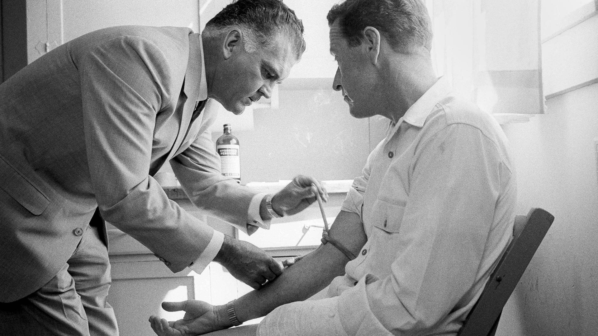 Historisches Schwarzweißfoto eines Freiwilligen mit eingegipstem Arm, dem in einer Klinikumgebung von einem Arzt LSD gespritzt wird.