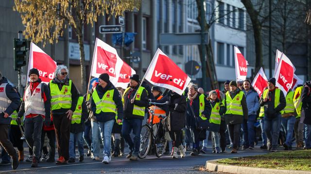 Demonstranten bei einem Warn-Streik. Die Menschen tragen gelbe Warn-Westen. Mehrere Menschen tragen Fahnen. Die Fahnen zeigen das Logo der Gewerkschaft Verdi.