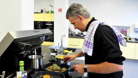 Jens Witt steht an einem Herd in einer Großraumküche und bereitet Essen zu. Er trägt ein Geschirrtuch über der Schulter. Jens Witt steht an einem Herd in einer Großraumküche und bereitet Essen zu. Er trägt ein Geschirrtuch über der Schulter.