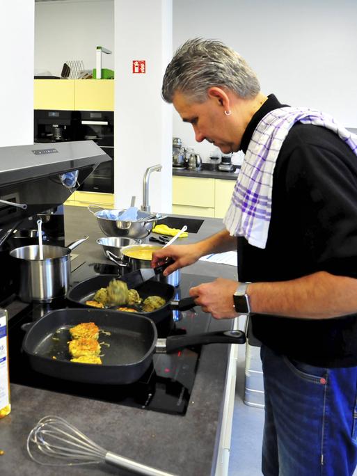 Jens Witt steht an einem Herd in einer Großraumküche und bereitet Essen zu. Er trägt ein Geschirrtuch über der Schulter.