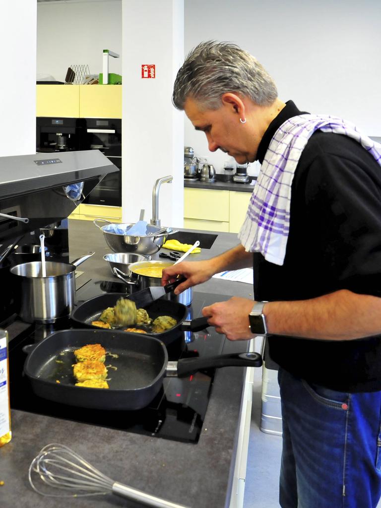 Jens Witt steht an einem Herd in einer Großraumküche und bereitet Essen zu. Er trägt ein Geschirrtuch über der Schulter.