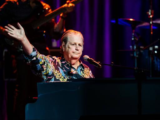 Brian Wilson sitzt in einem bunten Hemd am Konzertflügel, im Hintergrund die Band.