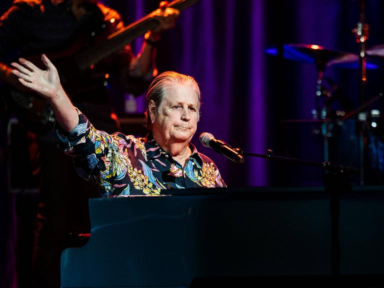Brian Wilson sitzt in einem bunten Hemd am Konzertflügel, im Hintergrund die Band.