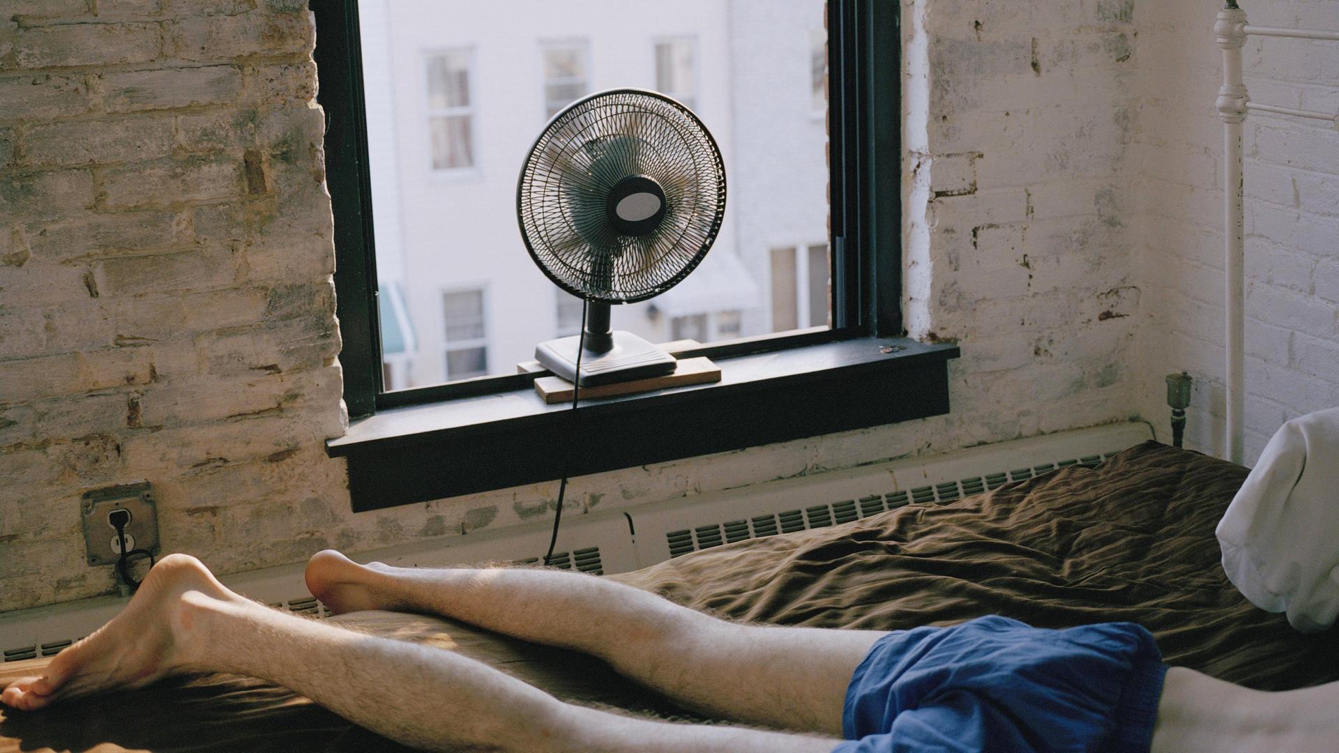 Ein Mann liegt in Unterhose auf einem Bett in einem Zimmer. Ein Ventilator steht auf dem Fensterbrett.