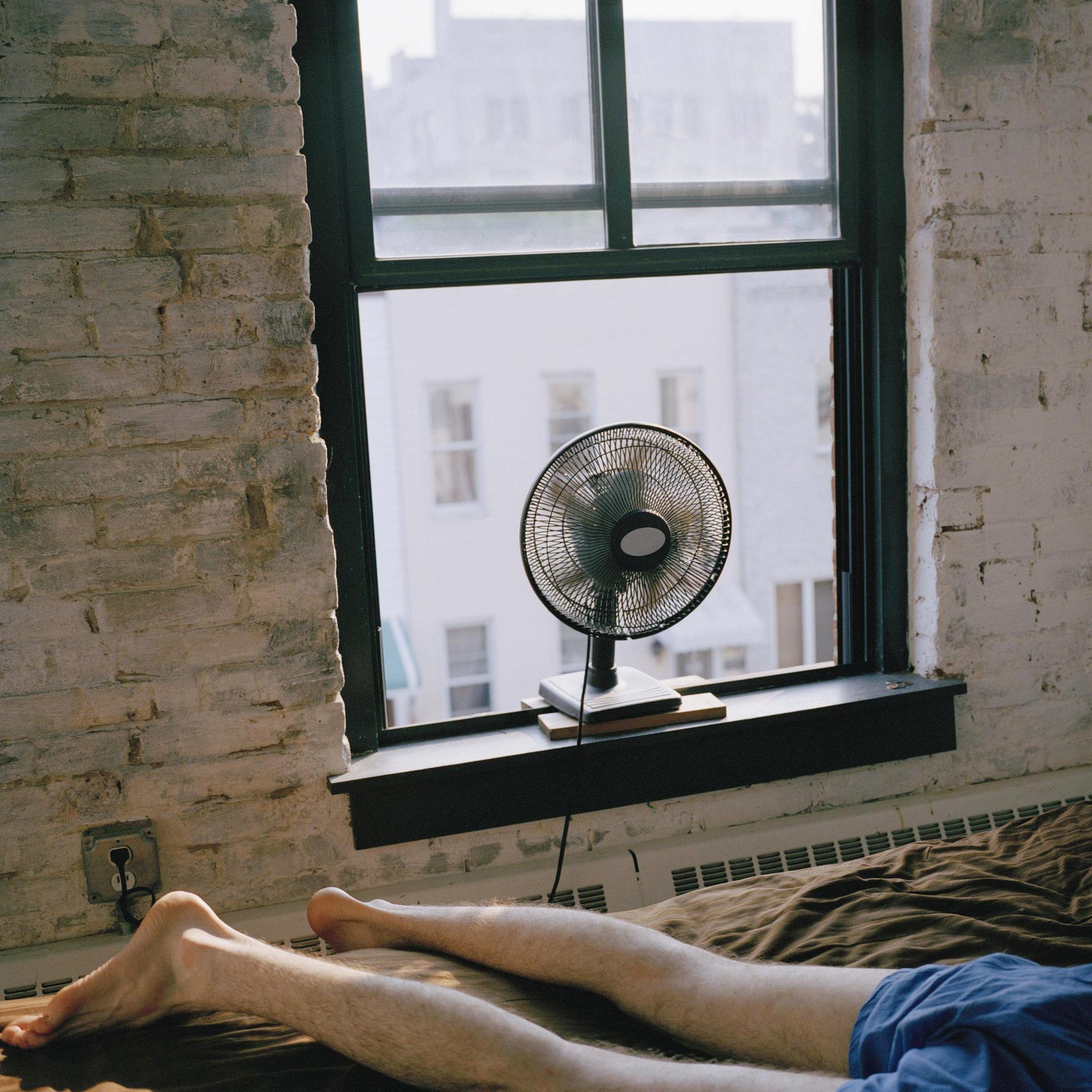 Ein Mann liegt in Unterhose auf einem Bett in einem Zimmer in New York City. Ein Ventilator steht auf dem Fensterbrett.
