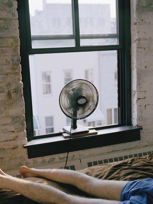 Ein Mann liegt in Unterhose auf einem Bett in einem Zimmer. Ein Ventilator steht auf dem Fensterbrett.