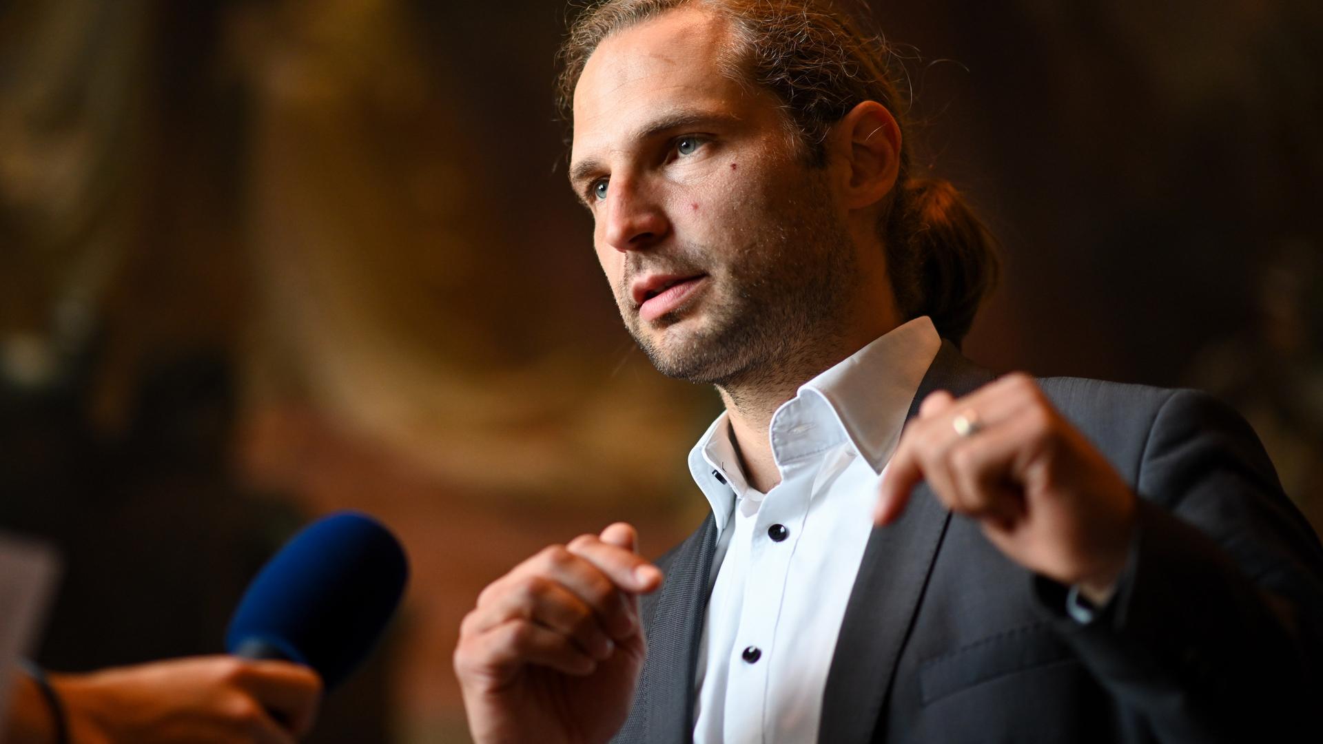 Toni Schuberl spricht während eines Interviews in ein Mikrofon. Er trägt einen Anzug und das blonde Haar im Nacken zum Zopf gebunden.