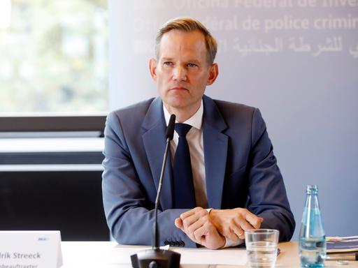 Ein Mann blick bei einer Pressekonferenz in die Kamera. Vor ihm ist ein Mikrofon. Er trägt einen dunklen Anzug, mit weißem Hemd und Kravatte. Es ist Hendrik Streeck, der Drogen- und Suchtbeauftragte der Bundesregierung.