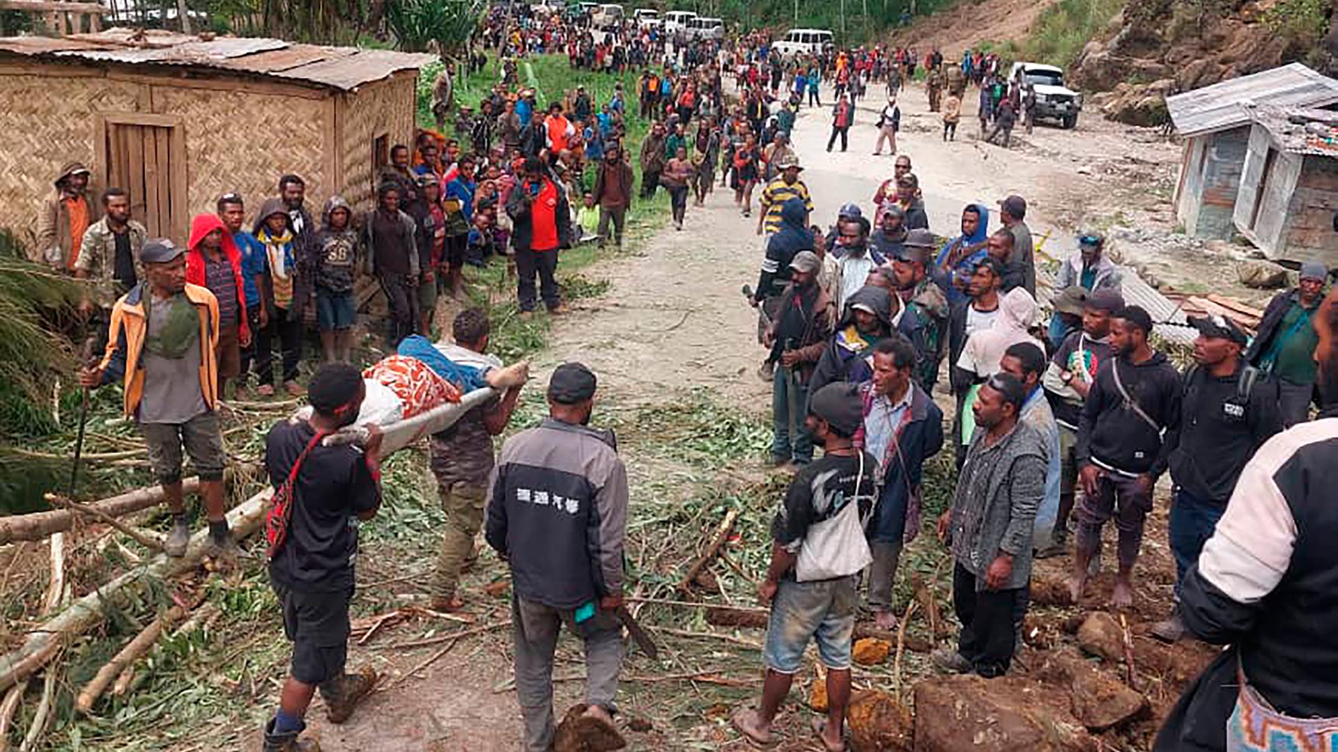 Eine verletzte Person wird nach dem Erdrutsch in Papua-Neuguinea zum Arzt getragen. Zwei Menschen haben die improvisierte Trage auf ihren Schultern, zahlreiche andere Menschen stehen am Wegrand. 