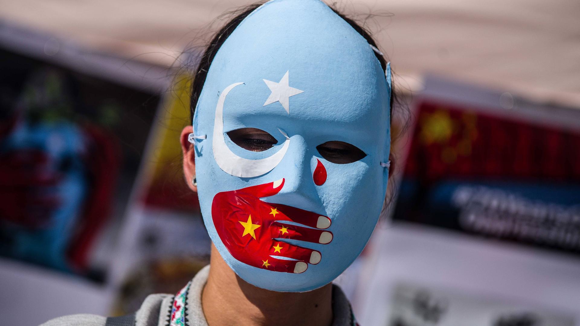 Eine Frau trägt eine blaue Maske mit weißem Stern und weißer Mondsichel. Über den Mund ist eine rote Hand gemalt, die eine chinesische Fahne symbolisiert. Im rechten Augenwinkel ist eine rote Träne. 