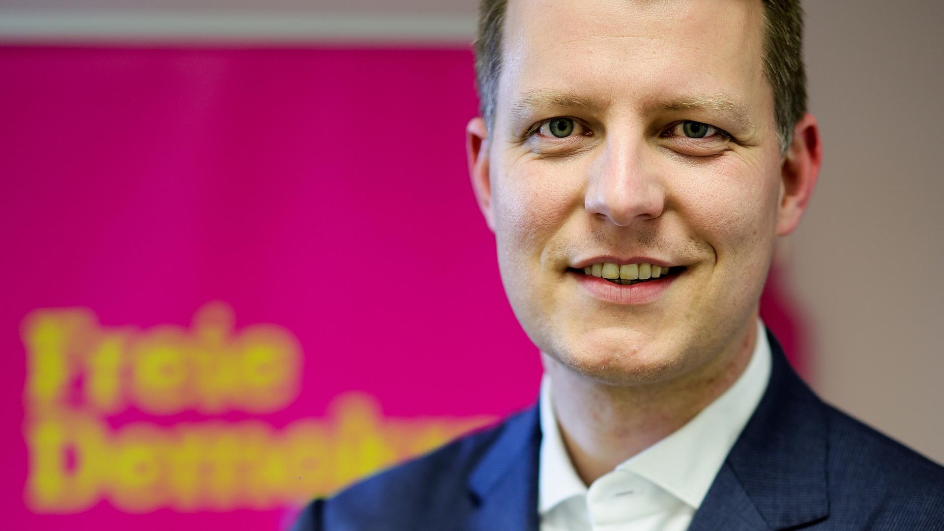 Henning Höne steht vor einem Plakat der Partei und schaut freundlich in die Kamera.