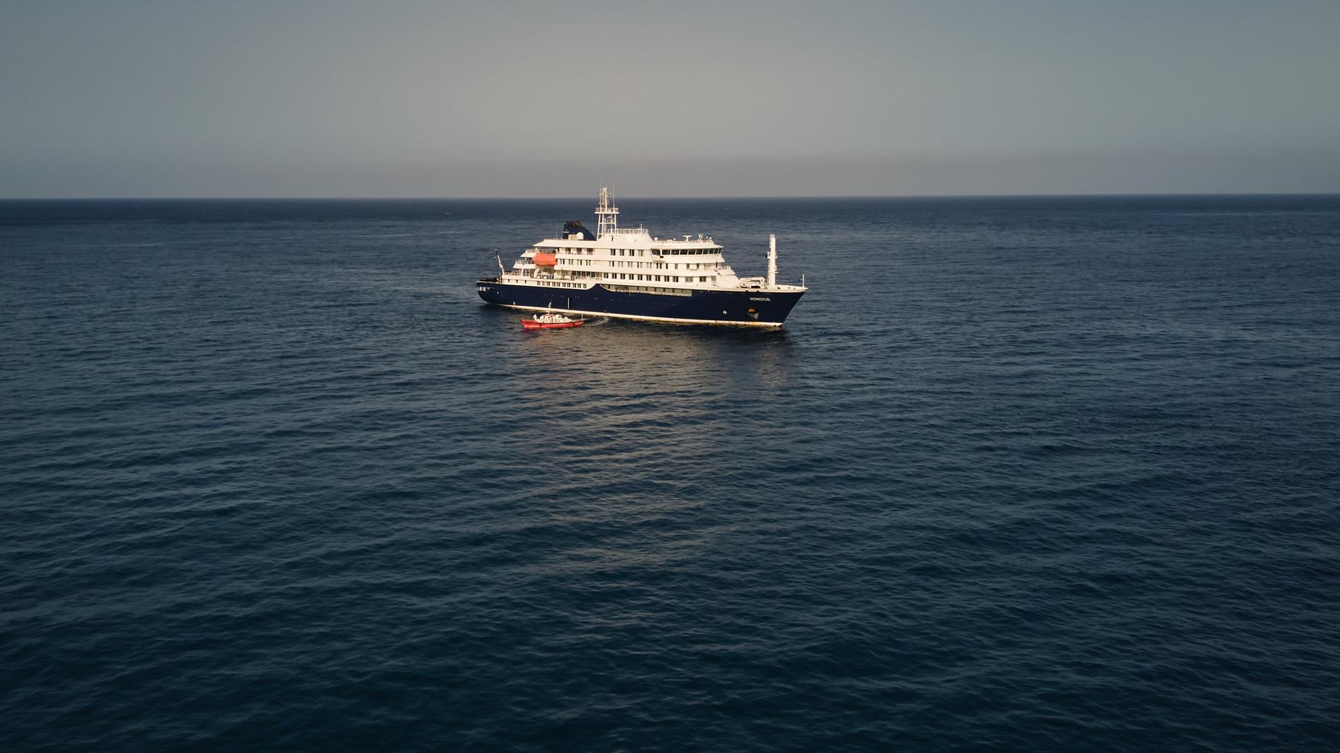 Auf der Luftaufnahme sieht man das kleine blau-weiße Schiff im dunkelblauen Meer. Am linken Bildrand ist das Ende einer Landzunge zu sehen. 
