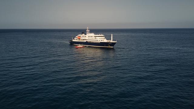 Auf der Luftaufnahme sieht man das kleine blau-weiße Kreuz-Fahrt-Schiff im dunkelblauen Meer. 
