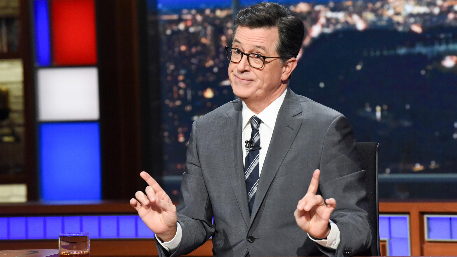 Der Showmaster Stephen Colbert sitzt während seiner Show an einem Schreibtisch mit Glas und Papier vor sich.