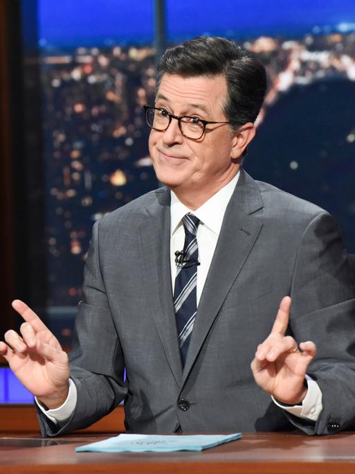 Der Showmaster Stephen Colbert sitzt während seiner Show an einem Schreibtisch mit Glas und Papier vor sich.