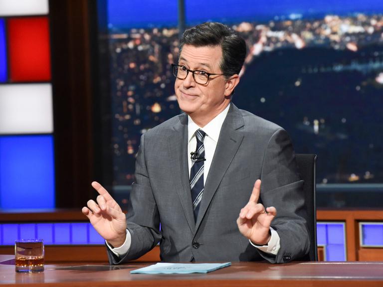 Der Showmaster Stephen Colbert sitzt während seiner Show an einem Schreibtisch mit Glas und Papier vor sich.