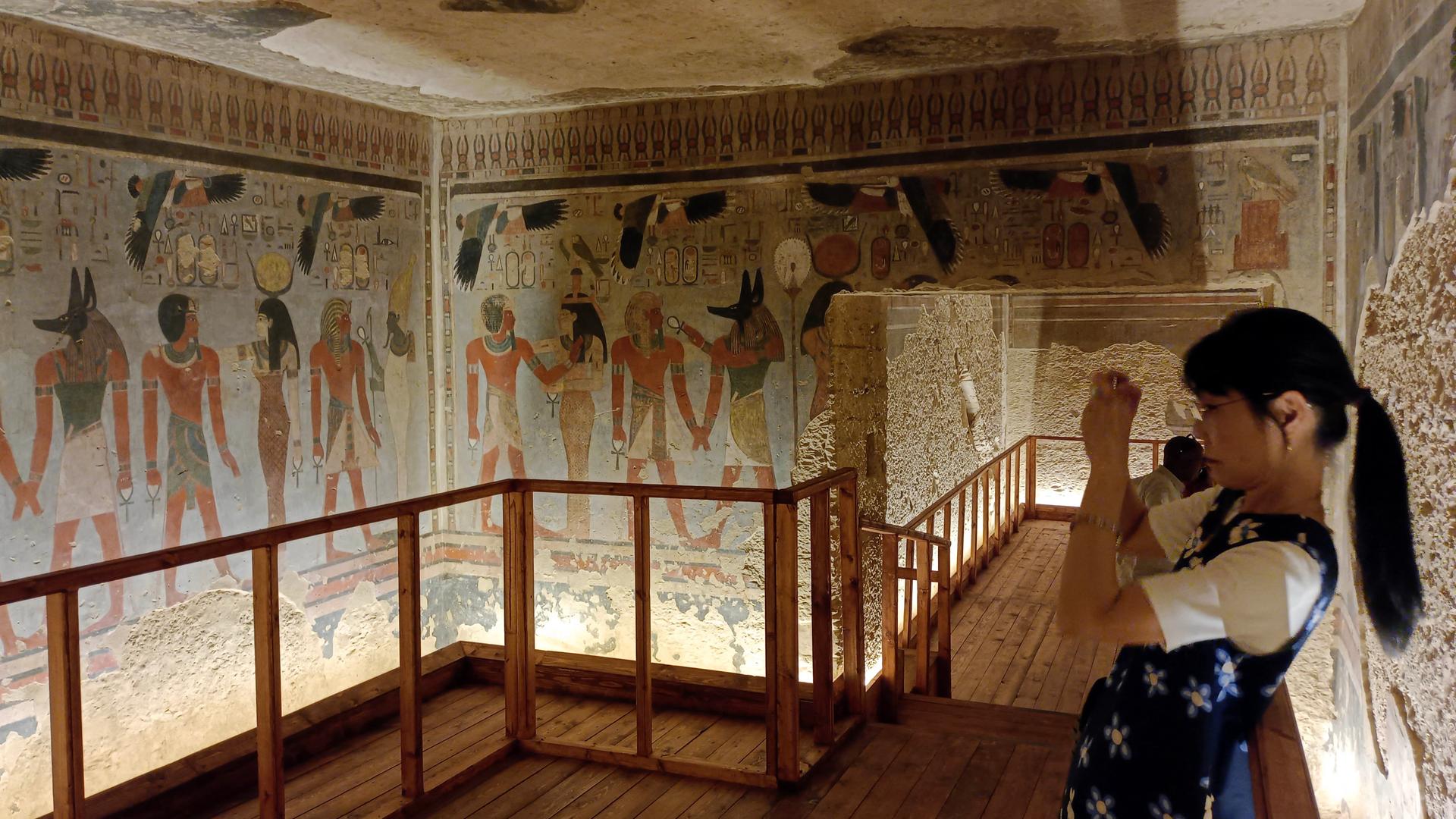 Eine Frau steht in der Grabkammer des Pharaos Amenhotep III. und macht ein Foto. Die Wand ist mit ägyptischen Fresken geschmückt.
