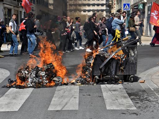 Eine brennende Mülltonne liegt auf der Straße, im Hintergrund ziehen Protestierende vorbei.