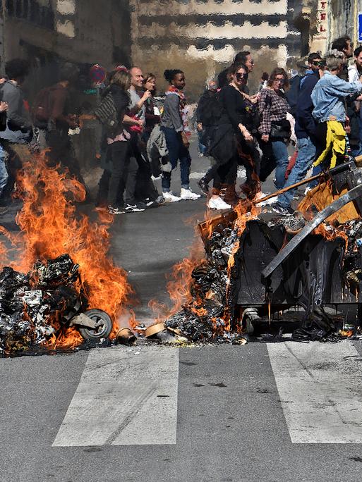 Eine brennende Mülltonne liegt auf der Straße, im Hintergrund ziehen Protestierende vorbei.