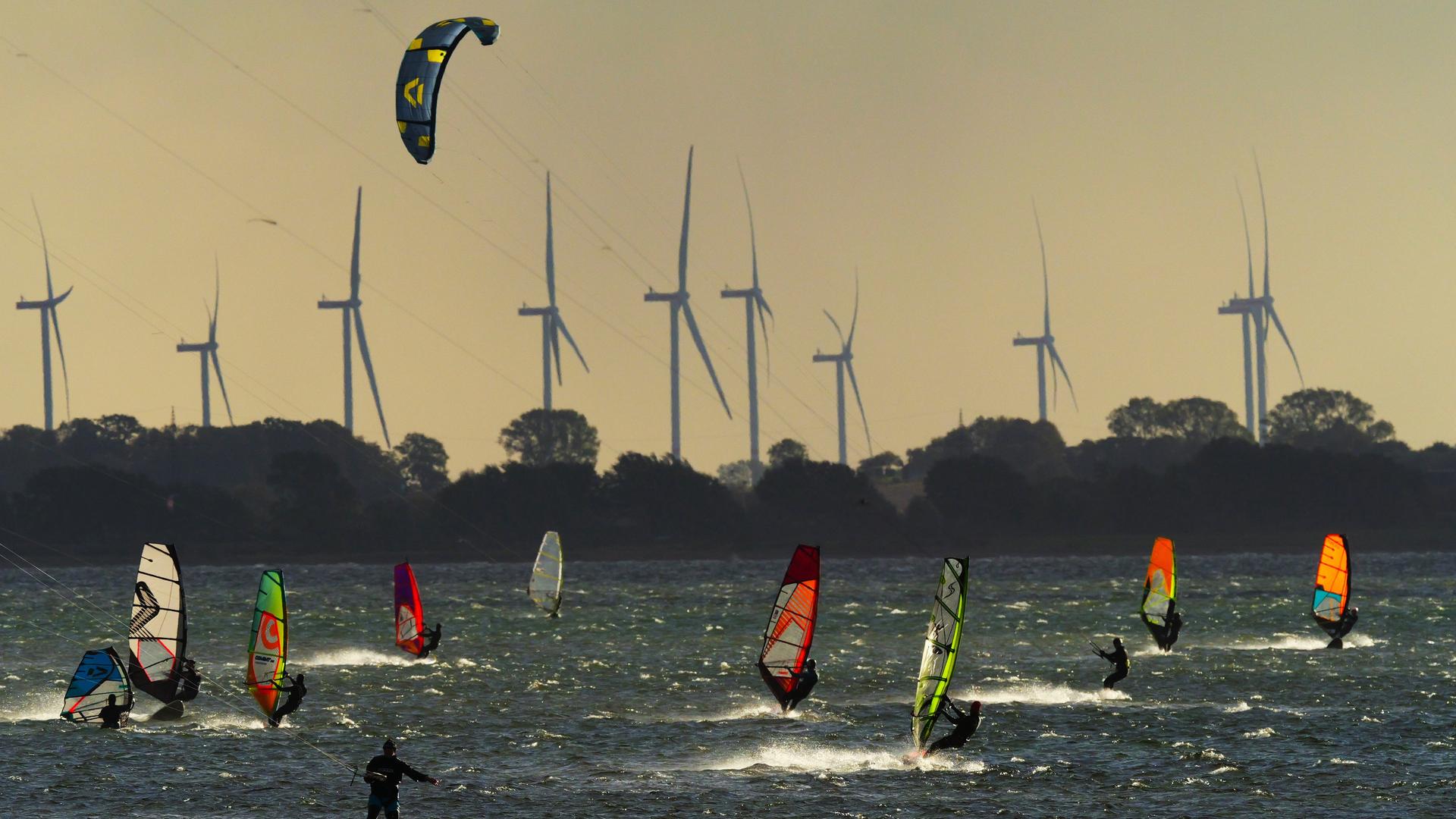 Surfer und und Kitesurfer an der Westküste von Fehmarn an der Ostsee. Im Hintergrund sind Windräder zu sehen. Vor dem kleinen Ort Gold auf Fehmarn liegt ein im weiten Umkreis beliebtes Surfrevier.