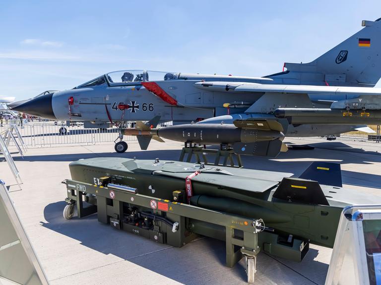 Im Vordergrund ein grüner Taurus-Marschflugkörper, im Hintergrund ein graues Kampfflugzeug der Bundeswehr