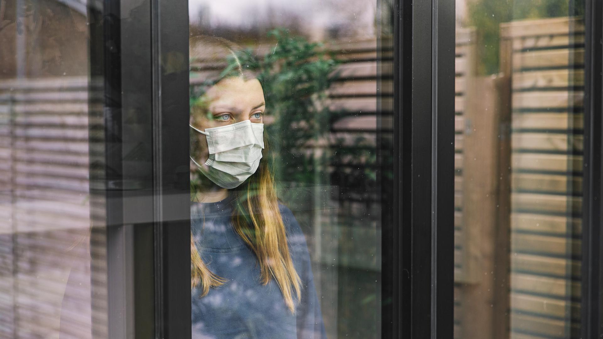 Eine junge Frau mit Maske schaut aus einem Fenster in dem sich ein Zaun und eine Pflanze spiegeln. 