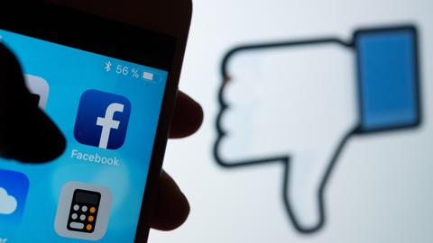 Das Logo des sozialen Netzwerks Facebook ist auf dem Display eines Smartphones zu sehen. Im Hintergrund wird auf einem Bildschirm ein das Zeichen für "Gefällt mir nicht" (Daumen runter) angezeigt.