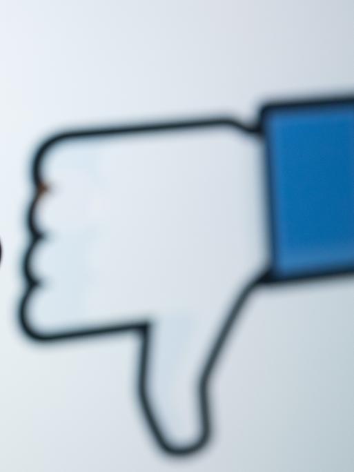 Das Logo des sozialen Netzwerks Facebook ist auf dem Display eines Smartphones zu sehen. Im Hintergrund wird auf einem Bildschirm ein das Zeichen für "Gefällt mir nicht" (Daumen runter) angezeigt.