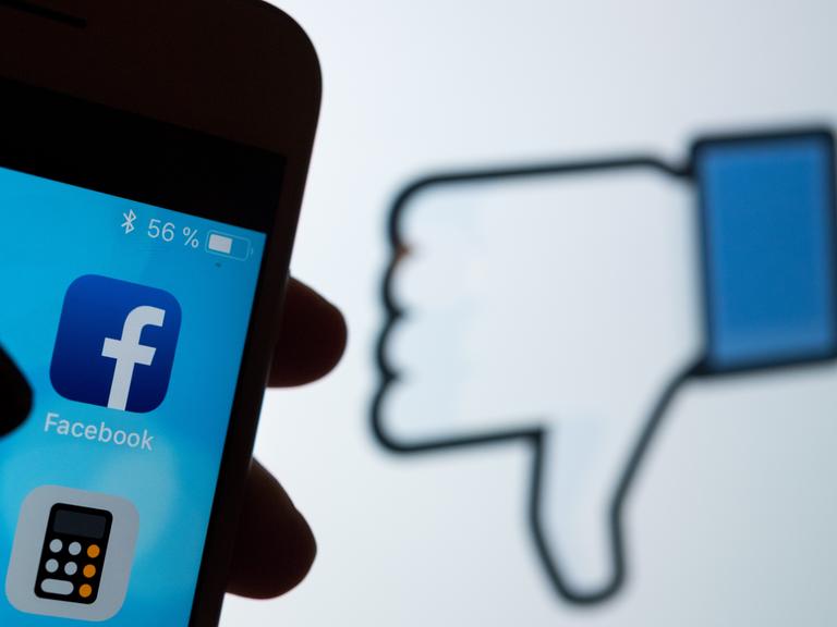 Das Logo des sozialen Netzwerks Facebook ist auf dem Display eines Smartphones zu sehen. Im Hintergrund wird auf einem Bildschirm ein das Zeichen für "Gefällt mir nicht" (Daumen runter) angezeigt.