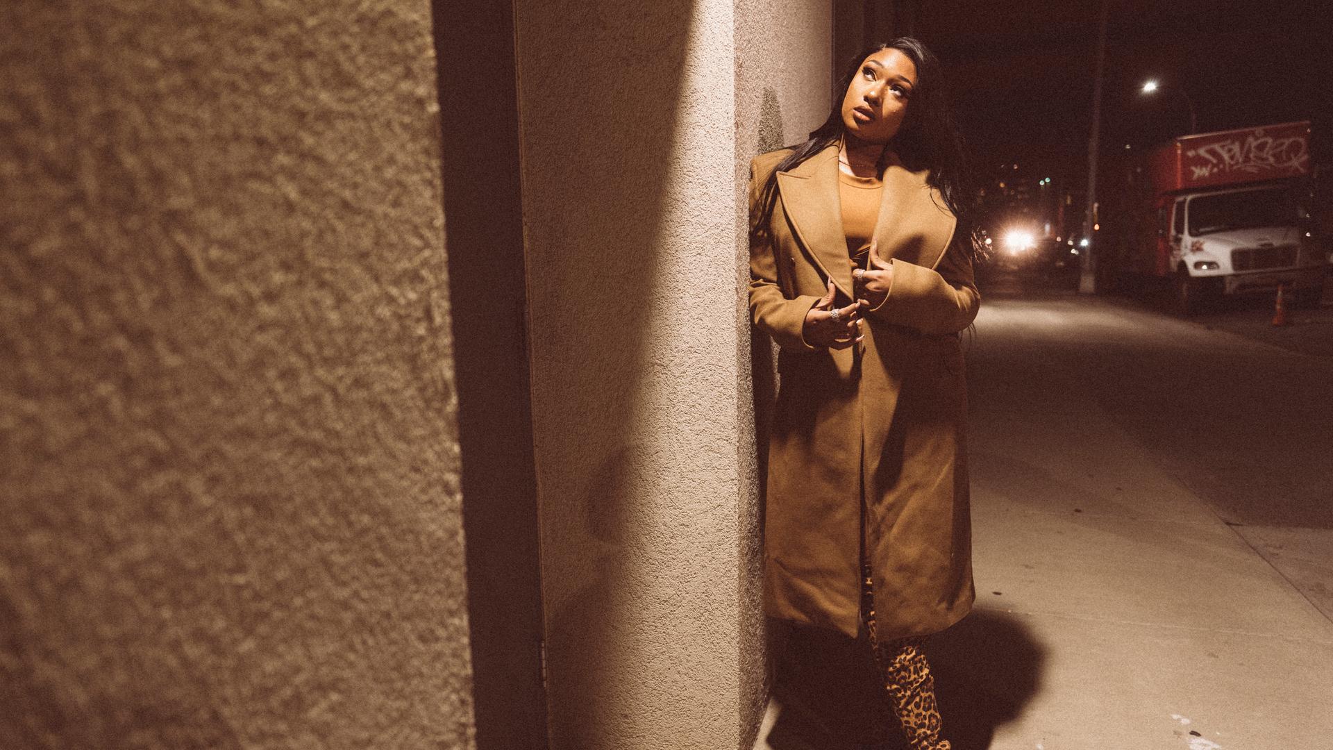 Die Rapperin Megan Thee Stallion steht mit Mantel und Leopardenstiefeln an einer dunklen Straßenecke. Die Rapperin Megan Thee Stallion steht mit Mantel und Leopardenstiefeln an einer dunklen Straßenecke.