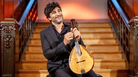Mandolinist Avi Avital sitzt mit seinem Instrument vor einer Holztreppe und lacht in die Kamera.