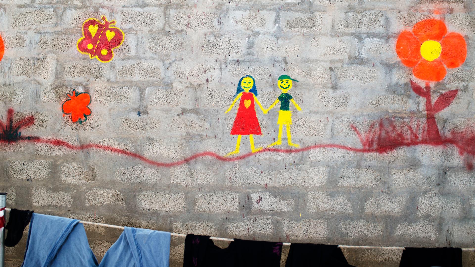 Ein Gemälde auf einer Backsteinwand im Lager Tampakika auf der Insel Chios zeigt ein Mädchen und einen Jungen, die sich an den Händen anfassen. Im Vordergrund hängt Wäsche an einer Leine. Ein Gemälde auf einer Backsteinwand im Lager Tampakika auf der Insel Chios zeigt ein Mädchen und einen Jungen, die sich an den Händen anfassen. Im Vordergrund hängt Wäsche an einer Leine.